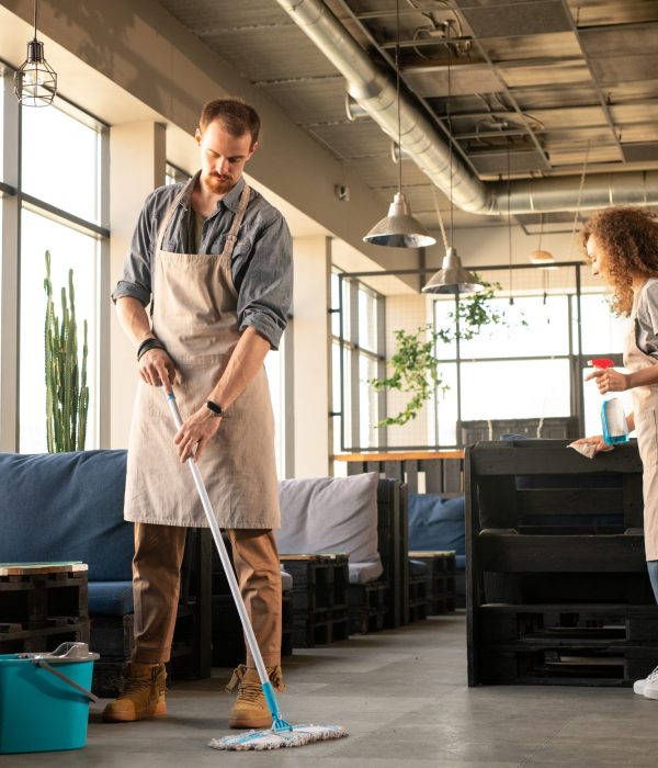 Busy young couple in aprons washing floor and cleaning surfaces with detergent while preparing cafe before opening